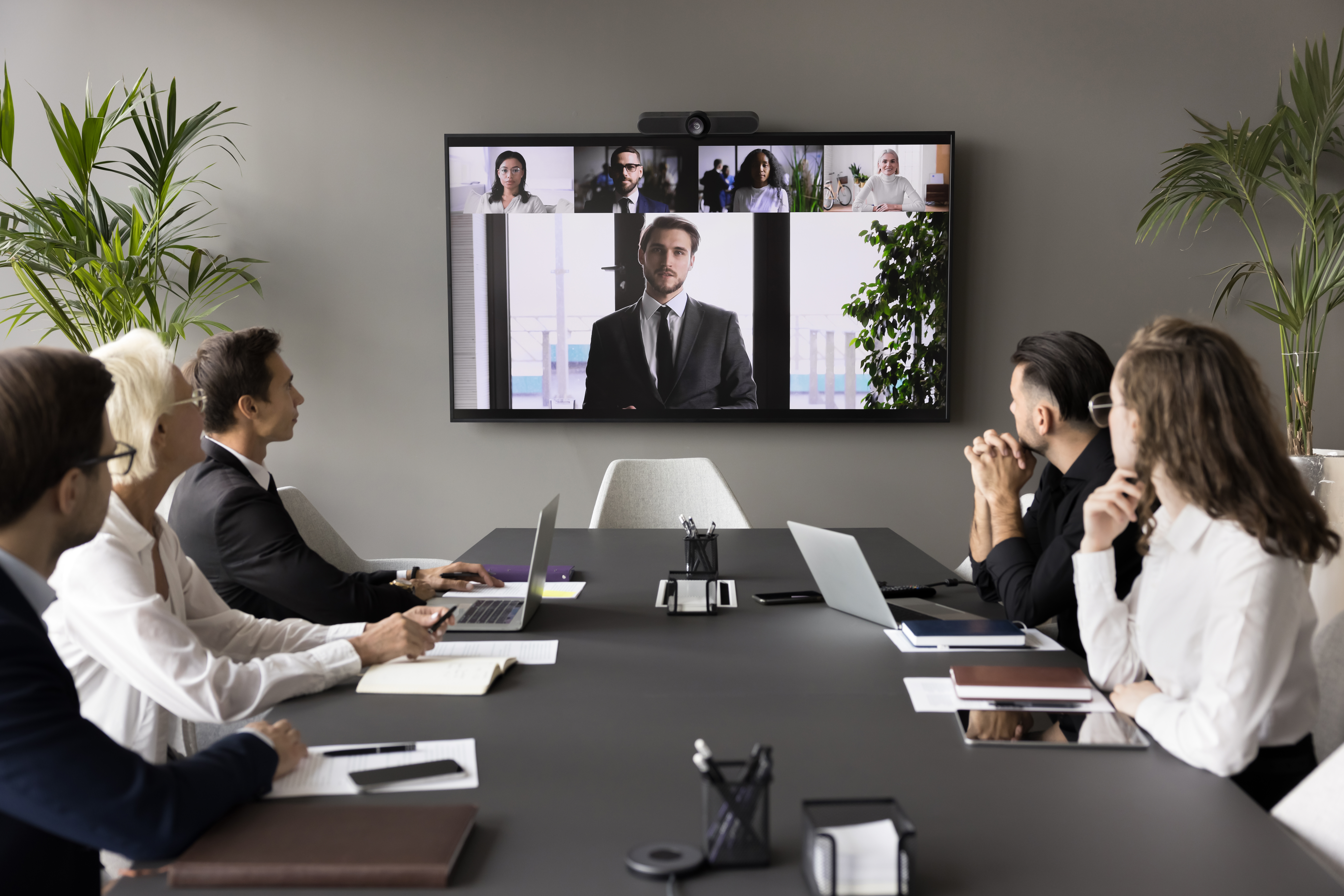 Business team and leader meeting online and offline, sitting at conference table, looking at interactive board with headshots, listening to speaker, CEO, discussing project on group video call